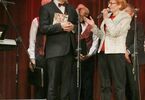 Kobieta trzymająca mikrofon i mężczyzna z kolorową torebką, za nimi mężczyźni w czerwonych kamizelkach