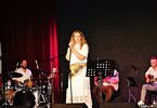 Zdjęcie: kobieta stojąca na scenie z mikrofonem, w tle mężczyźni z instrumentami