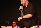 Zdjęcie: mężczyzna w ciemnej rozpiętej koszuli na scenie z mikrofonem, w tle osoby z instrumentami
