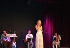 Zdjęcie: kobieta w długiej sukni z mikrofonem, w tle osoby z instrumentami