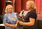 Dwie seniorki w kolorowych sukienkach. Jedna z nich trzymająca mikrofon i dyplom