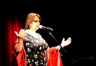 Kobieta w sukience w kwiaty stoi na scenie przed mikrofonem z rozłożonymi rękami