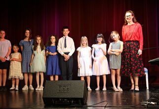 Zdjęcie: Uśmiechnięta kobieta i dzieci na scenie, w tle bordowa kotara