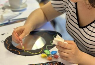 Zdjęcie: kobieta malująca czarną ramkę lusterka żółtymi kropkami farby, obok leżące ozdobione lusterko w technice dots