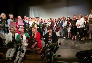 Zdjęcie: grupa osób na scenie, kobiety i mężczyźni, część z nich trzyma róże w ręku