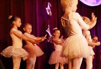 Zdjęcie: grupa dziewczynek w strojach baletowych w pozie tanecznej trzymająca ręce przed sobą, w tle bordowa kurtyna i wiszące pointy baletowe