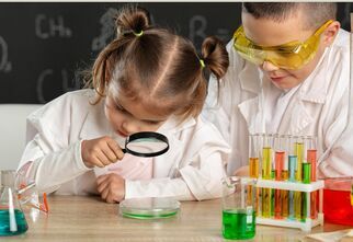 Zdjęcie: dziewczynka i chłopiec patrzący przez lupę, przed nimi probówki z kolorowymi cieczami