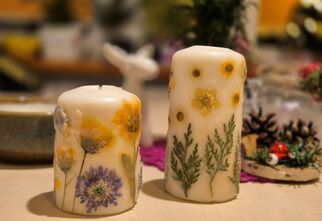 Zdjęcie: dwie białe świece z zatopionymi elementami kwiatów i ziół stojące na blacie