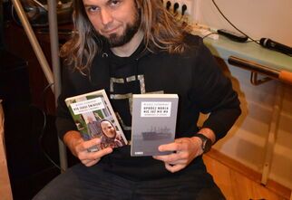 Zdjęcie: mężczyzna z długimi włosami siedzący przy stoliku trzymający w dłoniach dwie książki