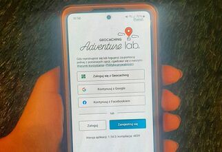 Zdjęcie: telefon komórkowy na tle zielonej trawy z widoczną aplikacją Adventure Lab trzymany w damskiej dłoni