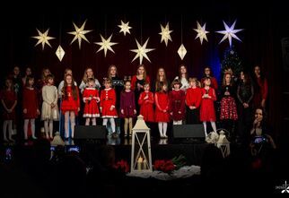 Zdjęcie: dzieci i młodzież na scenie w świątecznych stylizacjach, przed sceną kobieta z mikrofonem