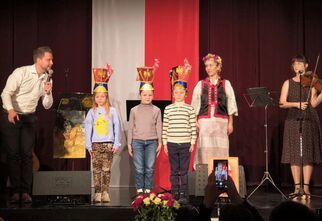 Zdjęcie: trójka dzieci w czapkach ułana stojąca na scenie, w tle biało-czerwona flaga, po prawej stronie młoda kobieta grająca na skrzypcach i stojąca obok w tradycyjnym stroju ludowym, po lewej stronie mężczyzna w białej koszuli z mikrofonem