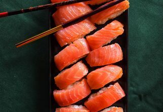 Zdjęcie: czarny prostokątny talerz z nigiri z łososiem na ciemnozielonym tle, na górze czarne pałeczki