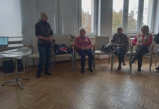 Zdjęcie stojącej starszej kobiety w otoczeniu seniorek siedzących na krzesłach