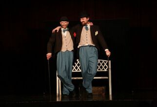 Zdjęcie: dwóch uśmiechniętych mężczyzn na scenie trzymających się za ramię, z tyłu biała ławeczka