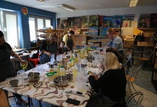 Zdjęcie: grupa kobiet w różnym wieku, stojące lub siedzące przy stole, widoczne stojące na nim garnki, butelki, miski, miarki, w tle ściana z obrazami