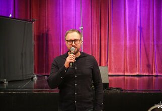 Zdjęcie: mężczyzna w okularach z mikrofonem w ciemnej koszuli, w tle kotara w fioletowo-różowym odcieniu