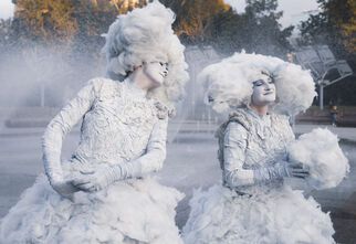 Zdjęcie: dwie osoby w białych, fantazyjnych kostiumach przypominających chmury lub pianę siedzące obok siebie, stroje i makijaż w odcieniach bieli. Jedna z postaci z delikatnym uśmiechem, w tle mgiełka lub woda z fontanny oraz drzewa.