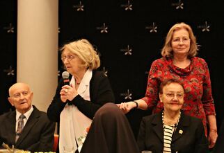 Zdjęcie: kobieta z mikrofonem i grupa osób siedzących przy stole w odświętnych strojach