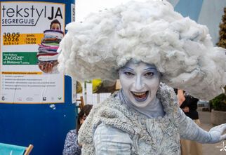 Zdjęcie: osoba w teatralnym kostiumie przypominającym chmurę, z białym makijażem i dużą puszystą peruką, za nią plakat na słupie z napisem Tekstylia segreguję