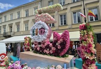 Zdjęcie: instalacja artystyczna z kwiatów, w jasnych kolorach, symbolizująca warszawską syrenkę, w tle kamienice