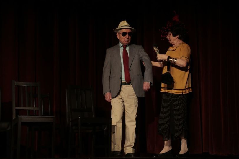 Mężczyzna w gariturze, kapeluszu i okularach stoi na scenie. Obok niego stoi kobieta i trzyma go za rękę na wysokości łokcia.