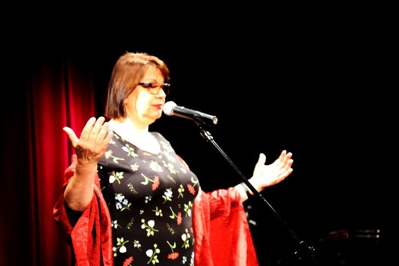Kobieta w sukience w kwiaty stoi na scenie przed mikrofonem z rozłożonymi rękami
