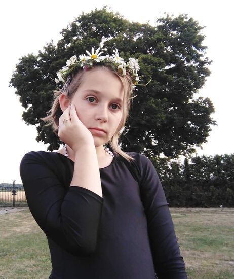 Dziewczynka trzymająca rękę na policzku. Na głowie ma wianek z polnych kwiatów. W tle wysokie drzewo.