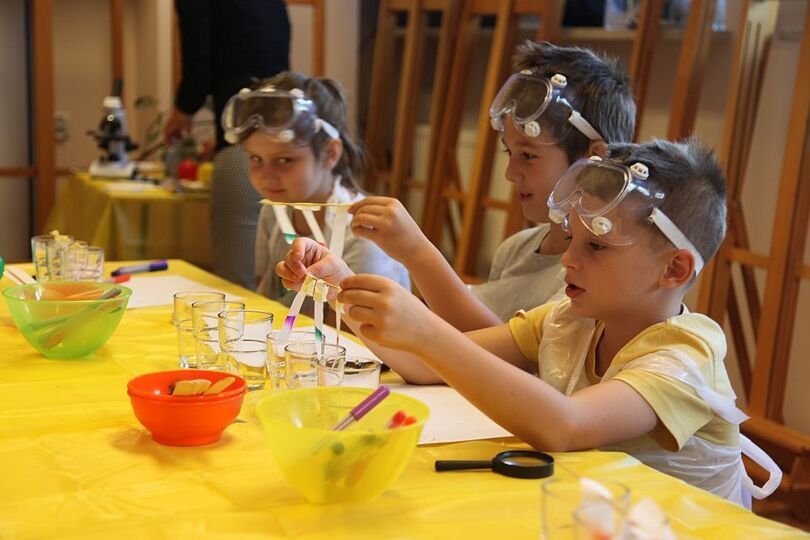 Dzieci siedzące przy stole z przedmiotami potrzebnymi do eksperymentów. Na głowach mają ochronne gogle.
