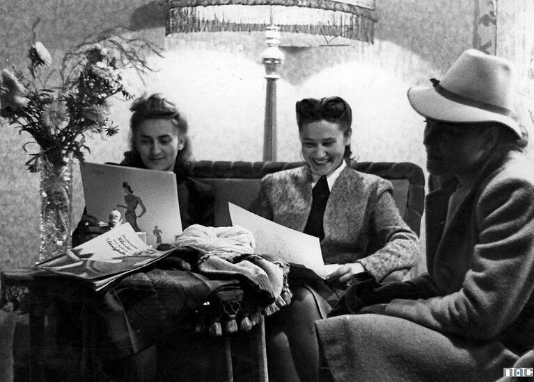 Zdjecie czarno-białe. Trzy eleganckie kobiety, jedna w kapeluszu, siedzą przy stole i oglądają katalogi mody