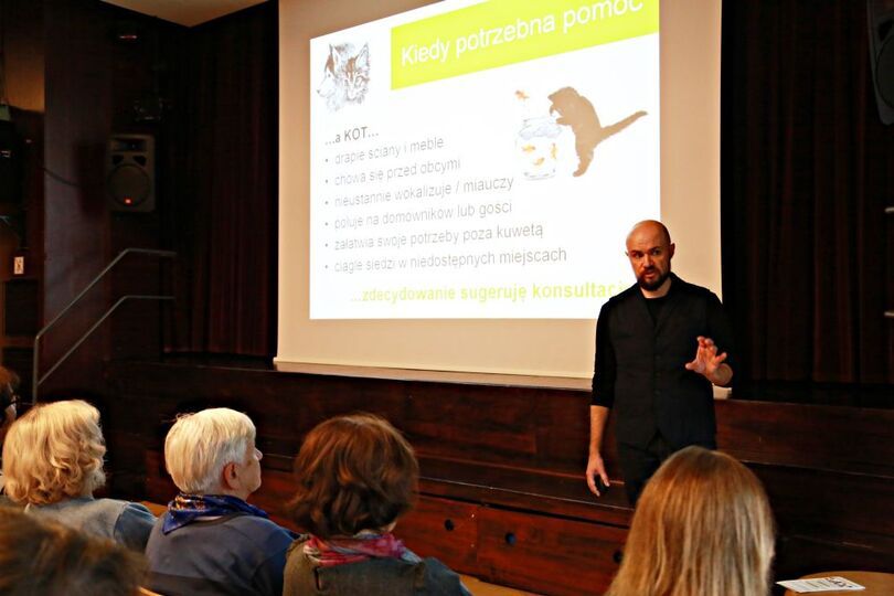 Gestykulujący behawiorysta podczas wykładu, za nim jeden ze slajdów prezentacji na temat tego, kiedy powinno się udać do behawiorysty z kotem