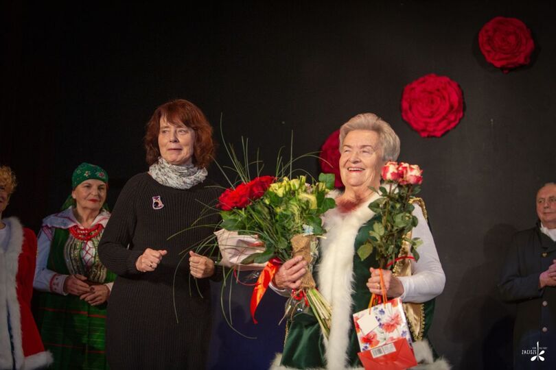Dwie kobiety, jedna w czarnej sukience, druga trzyma bukiet kwiatów i torebki prezentowe  i uśmiecha się