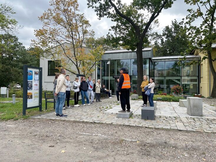 Grupa osób z przewodnikiem w pomarańczowej kamizelce, w tle szklany budynek