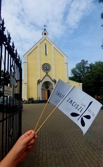 Dwie chorągiewki dom Kultury Zacisze na tle kościoła z żółtą elewacją