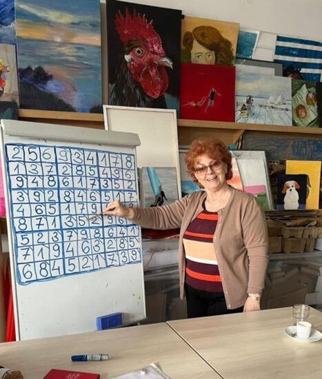 Zdjęcie: Uśmiechnięta kobieta-seniorka przy flipcharcie z grą sudoku, w tle kolorowe obrazy