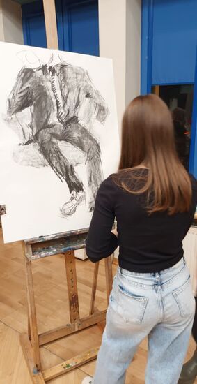 Zdjęcie: Młoda dziewczyna z długimi włosami stojąca przed rysunkiem postaci na sztaludze