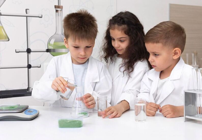 Zdjęcie: dziewczynka i dwóch chłopców w białych fartuchach, przelewający ciecz do szklanych probówek