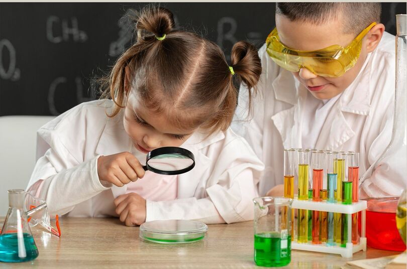 Zdjęcie: dziewczynka i chłopiec patrzący przez lupę, przed nimi probówki z kolorowymi cieczami