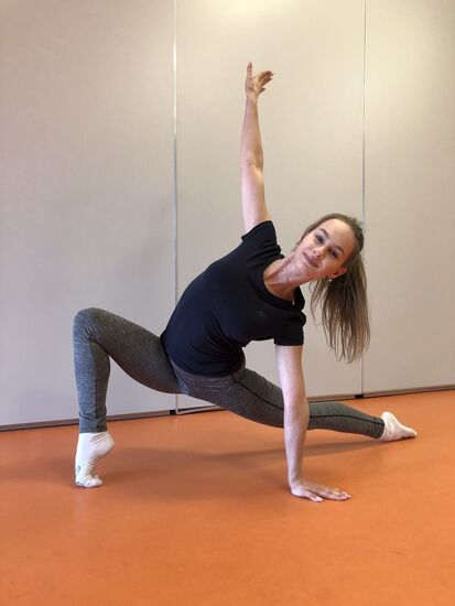 Zdjęcie: młoda dziewczyna ćwicząca w sali, w pozycji rozciągającej, jedna ręka oparta o podłogę, druga uniesiona w górę, nogi szeroko rozstawione