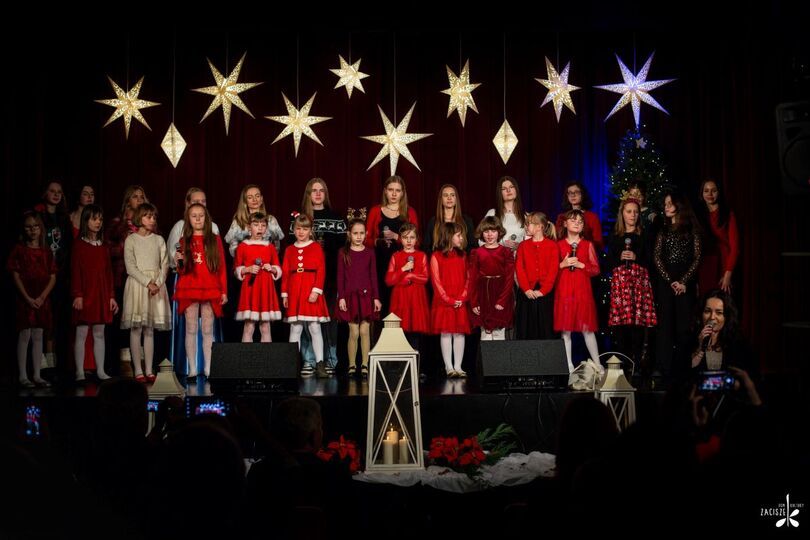 Zdjęcie: dzieci i młodzież na scenie w świątecznych stylizacjach, przed sceną kobieta z mikrofonem