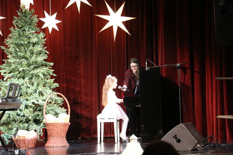 Zdjęcie: dziewczynka ubrana na biało siedząca na stołku przy pianinie, nad nią z mikrofonem stojąca kobieta w okularach, po lewej stronie choinka, pod nią dwa wiklinowe kosze z prezentami, na górze zawieszone gwiazdy