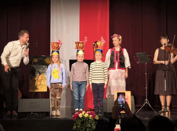 Zdjęcie: trójka dzieci w czapkach ułana stojąca na scenie, w tle biało-czerwona flaga, po prawej stronie młoda kobieta grająca na skrzypcach i stojąca obok w tradycyjnym stroju ludowym, po lewej stronie mężczyzna w białej koszuli z mikrofonem