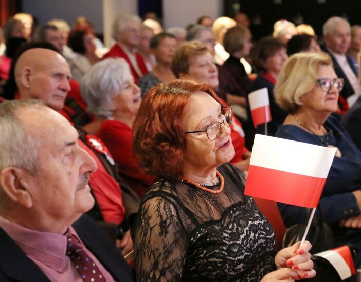 Zdjęcie: grupa seniorów w odświętnych strojach, na pierwszym planie seniorka z biało-czerwoną flagą