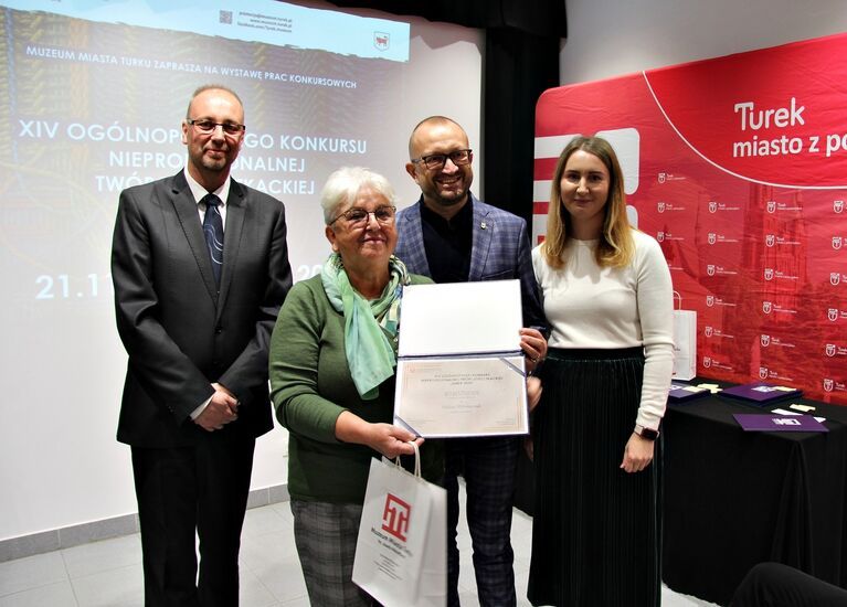 Zdjęcie: grupa uśmiechniętych, odświętnie ubranych osób, na pierwszym planie kobieta z otwartym dyplomem