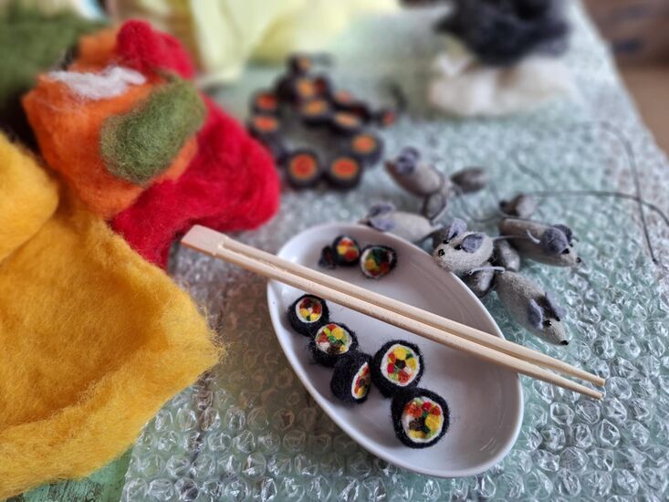 Zdjęcie: filcowe sushi w białym naczyniu, nad nimi drewniane pałeczki, z prawej strony kilka filcowych myszek, po lewej stronie kolorowy filc