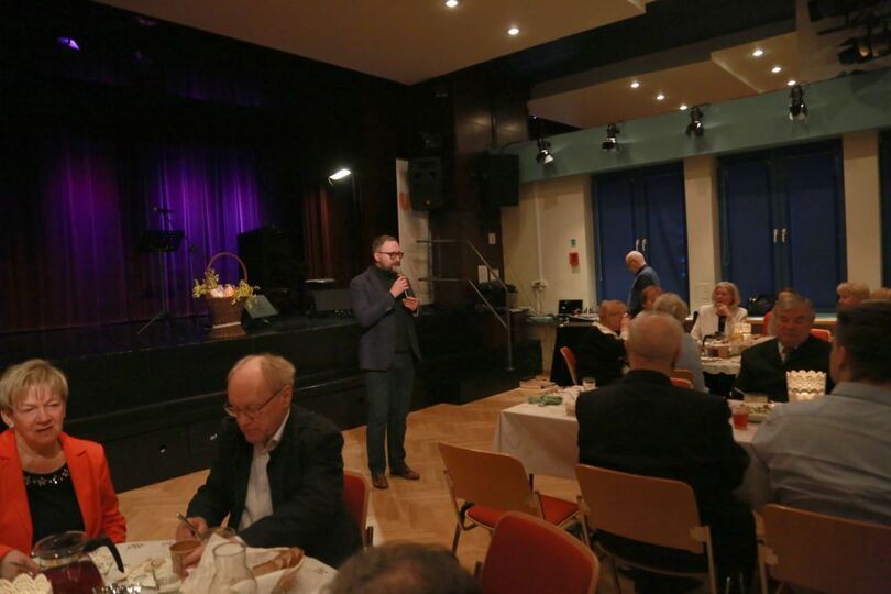 Zdjęcie mężczyzny trzymającego mikrofon, obok seniorzy siedzący przy stołach wielkanocnych