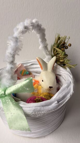 Zdjęcie: biały koszyczek z włóczki ozdobiony kokardą i suszonymi roślinami, w środku zajączek i pisanka