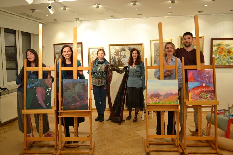 Zdjęcie: grupa czterech młodych osób stojących przy sztalugach z obrazami, z tyłu dwie kobiety przy harfie, w tle obrazy na ścianie