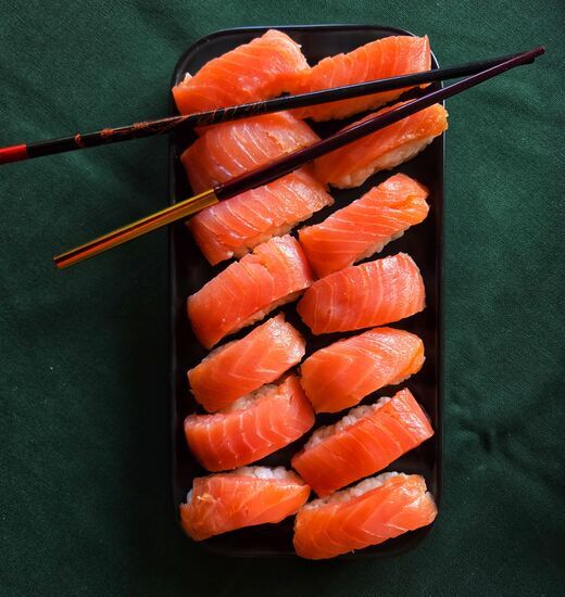 Zdjęcie: czarny prostokątny talerz z nigiri z łososiem na ciemnozielonym tle, na górze czarne pałeczki