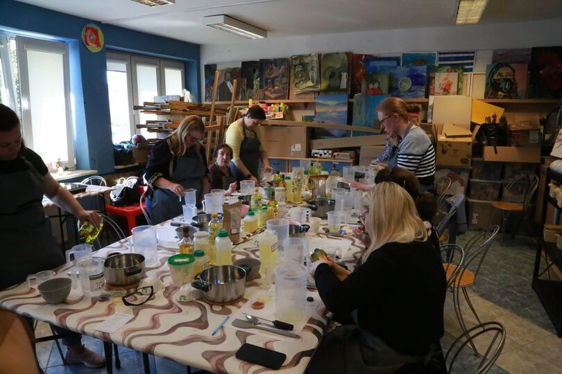 Zdjęcie: grupa kobiet w różnym wieku, stojące lub siedzące przy stole, widoczne stojące na nim garnki, butelki, miski, miarki, w tle ściana z obrazami